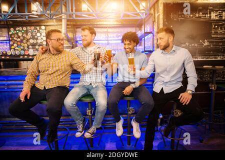 Vier fröhliche multikulturelle Freunde sitzen auf dem Barhocker und klirren mit Biergläsern vom Fass. Stockfoto