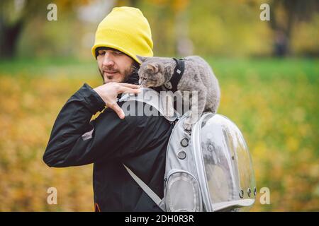 Street Walk Herbst, Erwachsene kaukasischen Mann trägt dunkle Jacke leuchtend gelb hut und seine trainierte inländische graue lustige Katze, männlich, sieht mit interest.the topi Stockfoto