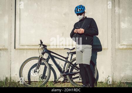 Der Kurier in Schutzmaske auf dem Fahrrad hielt an und beobachtete die Telefonanwendung. Bereitstellungsdienst in Quarantäne. Ein Mann Stockfoto