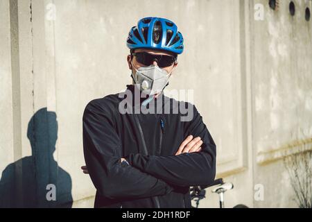 Ein Mann Radfahrer in einer schützenden Gesichtsmaske steht neben einem Fahrrad auf einem Hintergrund aus grauer Betonwand. Nicht erkennbarer Courier i Stockfoto