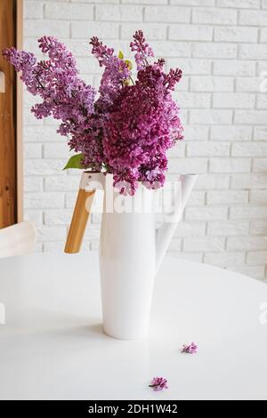 Bouquet von Flieder in einer weißen Gießkanne auf Weiß Tisch in der Nähe der Ziegelwand Stockfoto