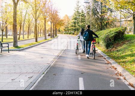 Zwei junge Braut und Bräutigam gehen auf einem Radweg Mit einem gemeinsamen E-Bike in den Händen Ein schöner Park mit vielen Bäumen bei su Stockfoto