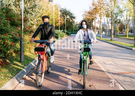 Vor zwei jungen Mann und Frau reiten entlang ein Fahrradweg mit einem gemeinsamen Elektro-Fahrrad in einem schönen park mit vielen Bäumen bei Sonnenuntergang tragen ein f Stockfoto