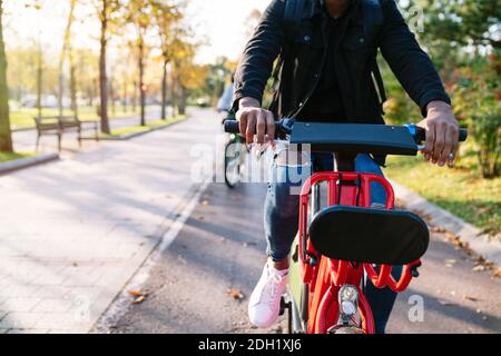 Nahaufnahme eines Jungen mit Rucksack Auf dem Rücken trägt er ein rotes Elektrofahrrad mit sich Ein von Bäumen gesäumter Parkweg bei Sonnenuntergang Stockfoto