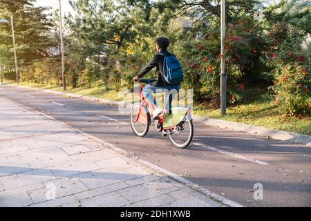 Junger Mann ein marokkanischer Student auf einem gemeinsamen E-Bike in einem schönen Park mit vielen Bäumen bei Sonnenuntergang, trägt eine Gesichtsmaske für die Coronavirus pand Stockfoto