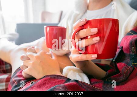 Rote Tasse in der Hand. Trinken heißen Tee oder Kakao. Verkürzte Seitenansicht von einem Mann und einer Frau, die Hände halten, die zu Hause auf einem Sofa im karierten Schlafanzug sitzen Stockfoto