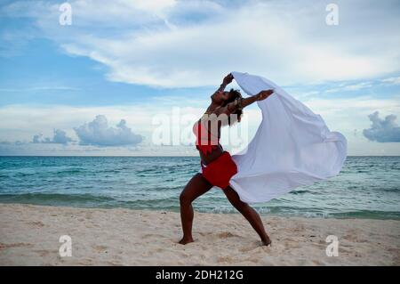 Die Frau tanzt mit rotem Kleid, um die Freiheit am Strand zu spüren. Eine junge Frau spielt mit dem Wind an einem Strand an der Karibik, Mexiko Stockfoto