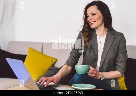 Erfolgreiche selbstbewusste Lehrerin mittleren Alters mit einer Tasse Kaffee vor dem Online-Unterricht, sitzend vor dem Laptop, mit Ele Stockfoto