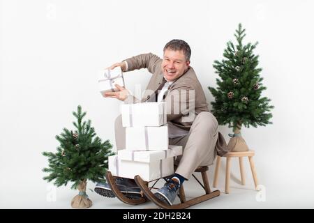 Weihnachten thematisierte horizontale Business-Porträt eines eleganten lässig gekleideten männlichen Executive sitzt auf einem Schlitten Jonglieren weiß verpackt Geschenkboxen alle auf einem weißen Hintergrund gesetzt. Stockfoto