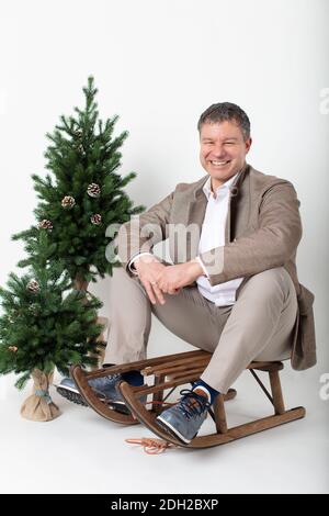 Weihnachten thematisierte horizontale Business-Porträt eines eleganten lässig gekleideten männlichen Executive sitzt auf einem rustikalen Holzschlitten auf einem weißen Hintergrund. Stockfoto