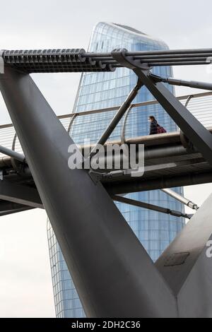 Aus dem unteren Winkel betrachtet eine Person mit Gesichtsmaske, die die Millennium Bridge mit einem Wolkenkratzer hinter sich überquert, London Stockfoto