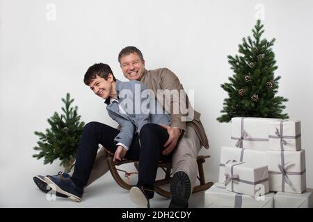 Weihnachten thematisierte horizontale Business-Paar Porträt gekleidet smart casual in blau, weiß, beige Hosen und Blazer auf grauem Hintergrund. Im Vordergrund ein Holzschlitten mit einem Stapel weißer Geschenkschachteln darauf. Zwei Miniatur-Weihnachtskiefern. Stockfoto