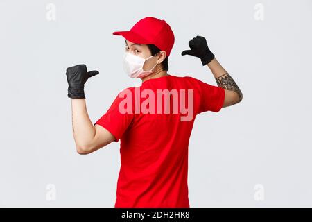 Rückansicht des asiatischen Lieferers in medizinischer Maske und Schutzhandschuhen, rote Kappe, T-Shirt, Kamera drehen, zeigt zurück zu s Stockfoto