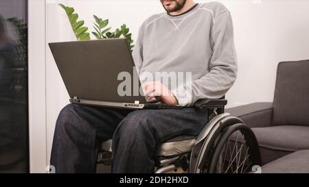 Junger Mann im Rollstuhl mit Laptop, der von zu Hause aus arbeitet, das Internet nutzt und auf den Bildschirm schaut. Stockfoto