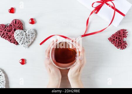 Frau Hände halten Tee Tasse in einem Herz mit roten Herzen Dekoration auf weißen Holztisch Draufsicht geformt. Konzept zum Valentinstag. Valentinstag Stockfoto