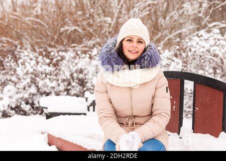 Ein junges Mädchen sitzt auf einer Bank in einem verschneiten Park. Es schneit im Winter. Mädchen in Winterkleidung in einem verschneiten Wald Stockfoto