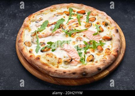 Meeresfrüchte-Pizza, Marinara, di Mare, mit Lachs, Muscheln und Rucola, auf einem Holzbrett, vor dunklem Hintergrund Stockfoto