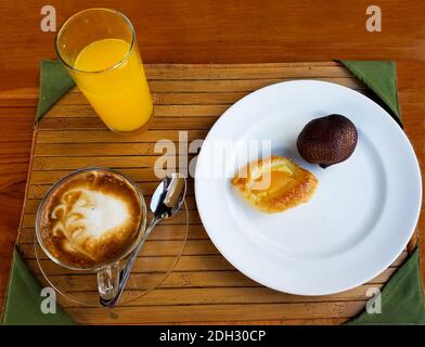 Minimalistisches asiatisches Frühstück mit einem Glas Orangensaft, Cappuccino-Kaffee und einem Teller mit einer Tarte und einer ungeöffneten Schlangenfrucht auf einem Bambustablett Stockfoto