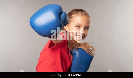 Fröhliches weibliches Kind in Boxhandschuhen, die Luft stanzen und lächeln Beim Stehen vor grauem Hintergrund Stockfoto