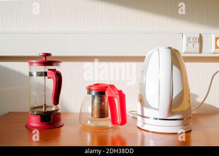 Eine französische Presse, ein Glaskessel und ein elektrischer Wasserkocher auf dem Tisch im Licht der Sonne. Küchenausstattung für die Zubereitung von Kaffee, Tee und anderen heißen Getränken. Stockfoto