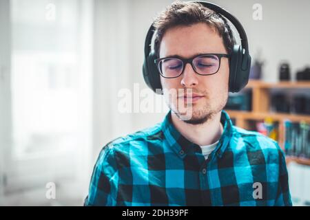 Musik zu Hause genießen: Der junge kaukasische Mann hört Musik mit Kopfhörern. Hemd mit blauem Karomuster. Stockfoto