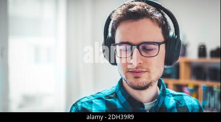 Musik zu Hause genießen: Der junge kaukasische Mann hört Musik mit Kopfhörern. Hemd mit blauem Karomuster. Stockfoto