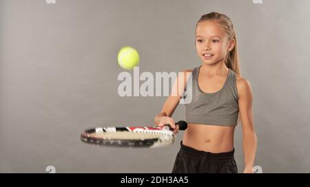 Nettes weibliches Kind in Sportkleidung, das Tennis spielt und lächelt, während Stehen vor grauem Hintergrund Stockfoto