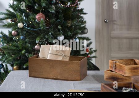 Eingewickelt in beigefarbenem Bastelpapier werden die gebundenen Geschenke in eine Holzkiste gefaltet. Sie liegen auf dem Tisch vor dem Hintergrund eines geschmückten Weihnachtsbaums. Stockfoto