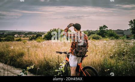 Ein junger Mann mit einem Fahrrad, der in den Himmel schaut. Ein junger Mann auf einem Fahrrad in der Natur, auf dem Feld schaut in die Ferne, ein Sportmann auf einem Fahrrad. Stockfoto
