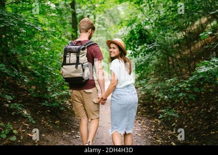 Glückliches kaukasisches Paar gehen in einem dichten Wald entlang des Pfades, Hände haltend, Rückansicht. Wanderer mit Rucksack auf der Suche nach p Stockfoto