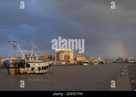 Ein geschwollener schlammiger Fluss Tiber fließt durch Fiumicino Hafen kurz nach einem schweren Sturm. Einige Fischerboote zurück von der Arbeit. Bläulicher Himmel. Sonnenstrahl Stockfoto