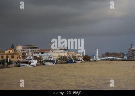 Ein geschwollener schlammiger Fluss Tiber fließt durch Fiumicino Hafen kurz nach einem schweren Sturm. Einige Fischerboote zurück von der Arbeit. Bläulicher Himmel. Sonnenstrahl Stockfoto