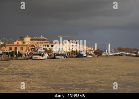 Ein geschwollener schlammiger Fluss Tiber fließt durch Fiumicino Hafen kurz nach einem schweren Sturm. Einige Fischerboote zurück von der Arbeit. Bläulicher Himmel. Sonnenstrahl Stockfoto