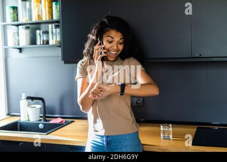 Junge hübsche Frau, die beim Sitzen an ihre Armbanduhr anschaut Auf einem Tisch in einer Küche Stockfoto