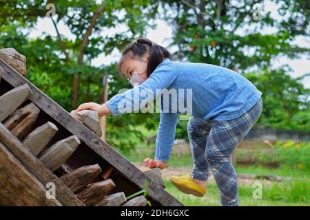 Ein süßes junges asiatisches Mädchen, das eine Maske trägt, klettert eine steile Holzrampe in einem Park hinauf, ergreift die Stufen mit ihren Händen und geht auf 4 Gliedmaßen. Stockfoto