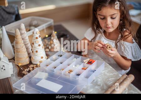 Nettes kleines Mädchen mit lockigen Haaren basteln und dekorieren Weihnachtsbaum Kegel mit Knöpfen. Stockfoto