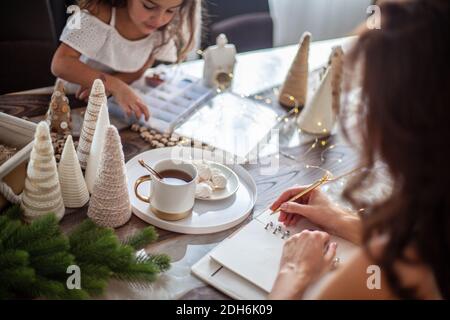 Junge Frau schreibt Pläne oder Ziele für das neue Jahr 2021 Während ihre Tochter Weihnachtsbäume basteln Stockfoto