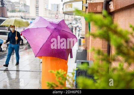 Frau in einem orangefarbenen Regenmantel mit einem lila Regenschirm bei Regenwetter auf dem Hintergrund der Stadt. Stadtbild bei schlechtem Wetter. Regentag. Stadtstraßenstil. Stockfoto