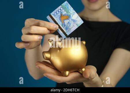 Frau in schwarzer Kleidung mit einer kasachischen Tenge und einem Sparschwein in den Händen. Stockfoto