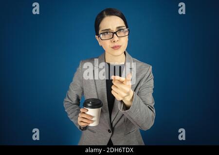 Ernst junge schöne Frau in einer grauen Jacke und Brille mit einem Pappbecher in der Hand auf dem blauen Hintergrund. Stockfoto