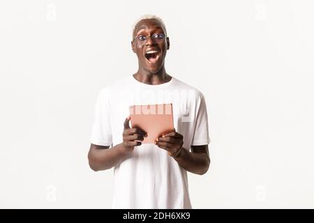Portrait von aufgeregt gut aussehend afroamerikanischen männlichen Schüler in Brille, Blick erstaunt, halten digitale Tablette, stehend weiß BA Stockfoto