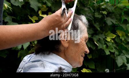 Schneiden Haare von älteren Frau zu Hause im Hinterhof im Freien. Zu Hause bleiben während der Quarantäne. Weibliche Hand hält Kamm und schneiden Haare mit einer Schere. Haarschnitt o Stockfoto