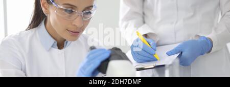 Junge Frau in weißer Uniform trägt schützende chemische Schutzbrille und Gummihandschuhe blicken ins Mikroskop Stockfoto