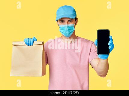 Delivery Mann halten Papierpaket mit Essen und Smartphone tragen Gesichtsmaske und Handschuhe als Schutz für COVID-19 Stockfoto