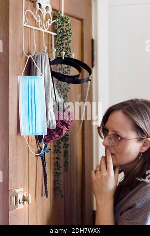 Eine Frau entscheidet, welche Schutzausrüstung sie vor dem Verlassen des Hauses tragen soll. Einweg-Gesichtsmaske, Stoffmaske, Hals-Buff und Gesichtsschutz hängen hinter der Tür Stockfoto