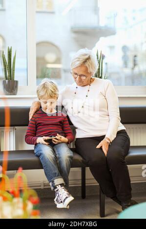 Großmutter und Enkel benutzen Smartphone, während sie in der Wartehalle sitzen Zimmer der orthopädischen Klinik Stockfoto