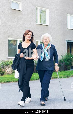 Porträt einer älteren Frau, die beim Gehen mit der Enkelin einen Stock hält Auf Fußweg gegen Gebäude Stockfoto