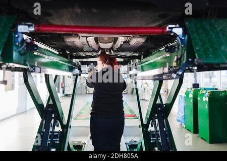 Rückansicht der Mechanikerin, die unter dem Fahrzeug auf der Hydraulik steht Aufzug Stockfoto