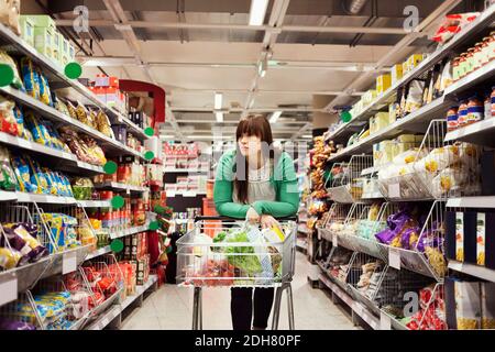 Junge Frau, die sich am Supermarkt-Gang auf den Einkaufswagen stützt Stockfoto