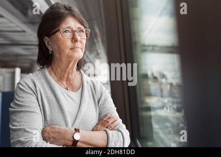 Durchdachte Geschäftsfrau, die im Büro Arme durch das Fenster gekreuzt hat Stockfoto